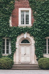 A beautiful house with a white door and windows, covered in ivy. The front of the building is seen from the outside,with green plants on both sides.A large red brick wall behind it forms an arch shape