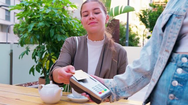 A woman is making a contactless payment using her card at a point-of-sale terminal in an outdoor setting. The scene shows a modern and easy way of paying, enhancing convenience and speed.