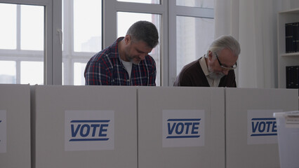 Concentrated men read ballot papers carefully while standing in voting booths during the...