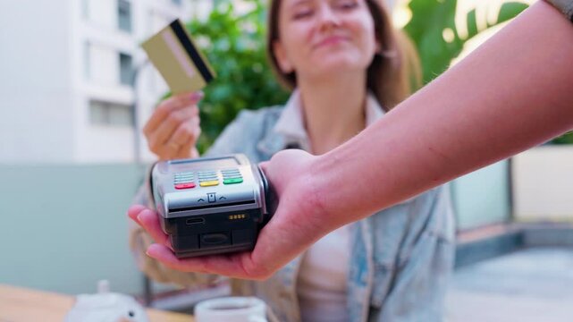 A woman is making a contactless payment using her card at a point-of-sale terminal in an outdoor setting. The scene shows a modern and easy way of paying, enhancing convenience and speed.