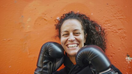 A smiling woman is posing against an orange wall, wearing boxing gloves