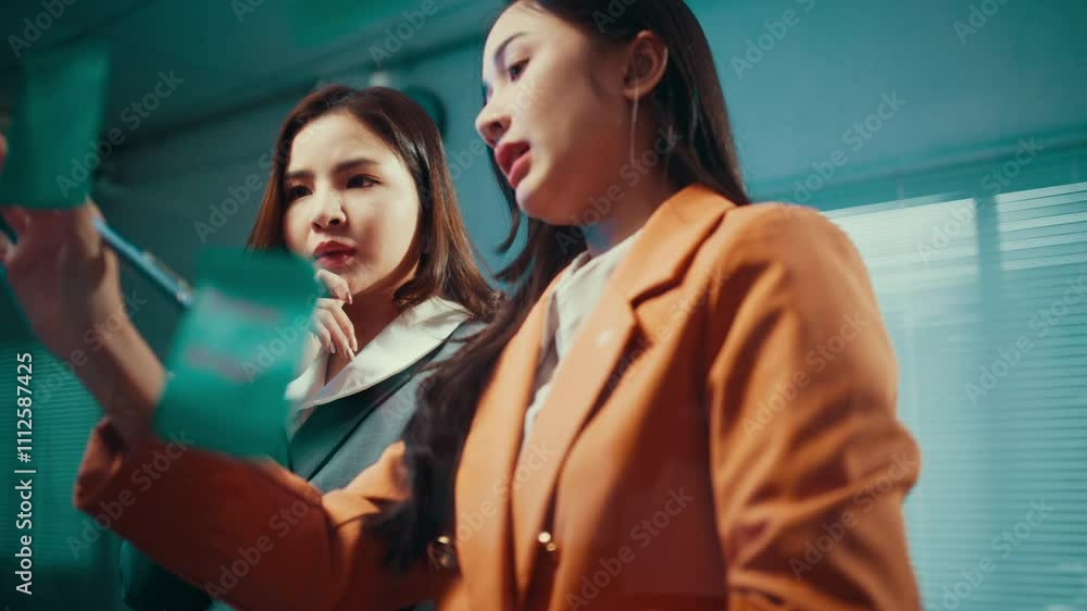 Poster two businesswomen brainstorm while using sticky notes on a glass wall in their office, fostering col