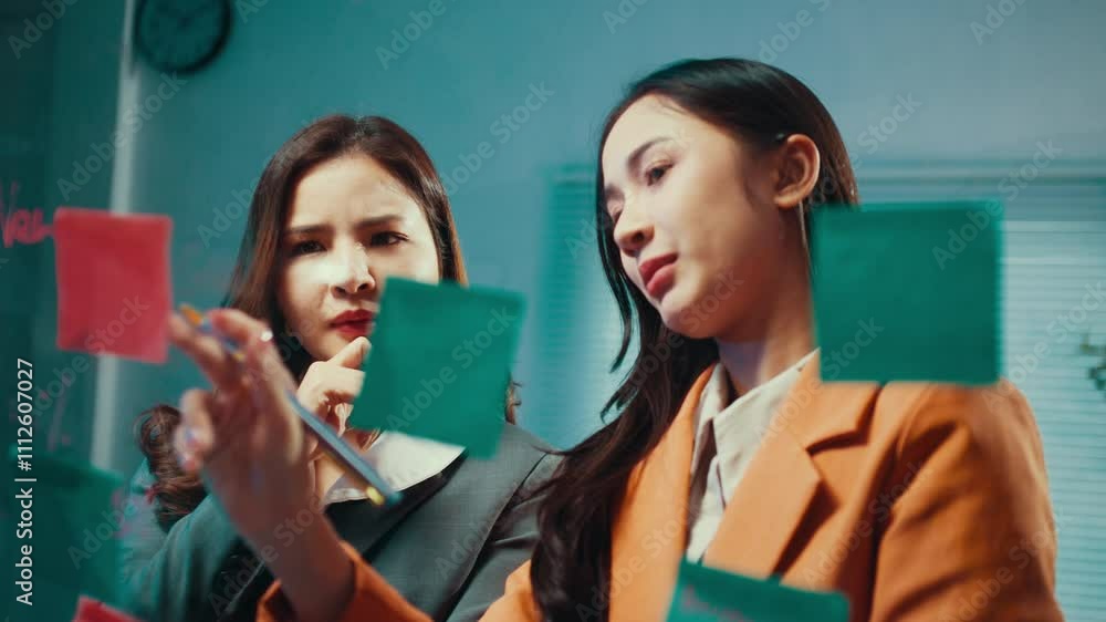 Poster two businesswomen engage in a discussion while utilizing sticky notes on a glass wall, fostering col