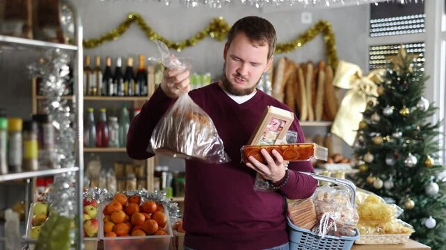 Young bearded man with basket full of goodies for Christmas celebration picking out traditional sweets in festively decorated shop, choosing between colorful panellets, rich turron and fresh panettone