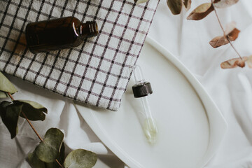  Pipette from a brown cosmetic jar for serum close-up on a white saucer with a towel and eucalyptus