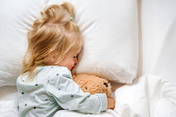 Little toddler girl child hugs a toy bear lying under a blanket, getting ready for bed