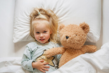 Little toddler girl child hugs a toy bear lying under a blanket, getting ready for bed