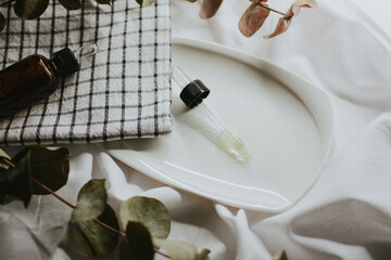  Pipette from a brown cosmetic jar for serum close-up on a white saucer with a towel and eucalyptus