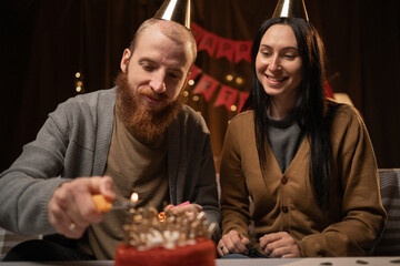 Young couple in party hats sitting near table and man lightning candles on birthday cake, celebrating together at home. family enjoying domestic anniversary party