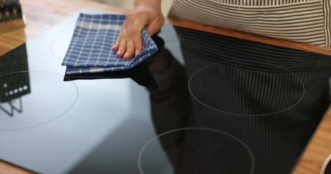 Woman cleans cooking surface of induction stove with checkered cloth in kitchen. Housewife keeps domestic appliance cleanness at home