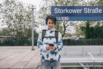 Teenage girl using smartphone while waiting for public transportation while listening to music on her headphones