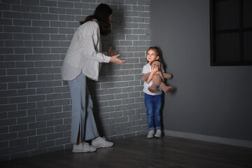 Sad little girl with toy bear and her angry mother standing in corner at home. Domestic violence...