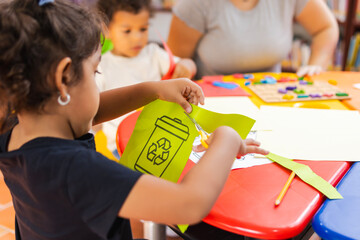Latina girl cutting out a paper with a drawing of a recycling bin, surrounded by classmates and...