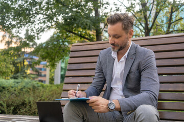 A confident business man, seen from the waist up, writes on his notepad while sitting on a park...