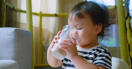 Adorable Cute Toddler child enjoying Drinking Chocolate Milk from a glass sitting in a cozy home,...