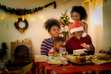 A joyful family gathers around the dinner table celebrating Christmas. The father, dressed in a Santa hat, gives a gift to the child, while everyone smiles with warmth and festive decorations.