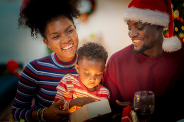 A heartwarming Christmas moment as a young child opens a beautifully wrapped gift with the help of his parents. The mother gently guides the child while the father, dressed in a Santa hat.