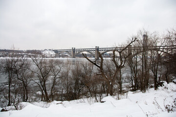 A picturesque view of a partially frozen river with a bridge in the distance, surrounded by snow and bare trees. Ideal for winter and travel-related content.