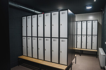 A well lit locker room features sleek metallic lockers with ventilation slots and secure locks, mounted above wooden benches. Dark walls and visible ductwork add an industrial touch.