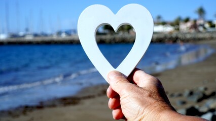 Valentines day seaside. Hand holds white painted wooden heart in front of the sea. Sailboats on horizon in the blurred background. Love and romance concepts