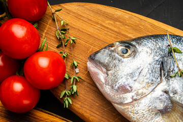 Raw dorado fish on a wooden board with oregano and cherry tomatoes on a black background, ready to cook. Close-up, top view.