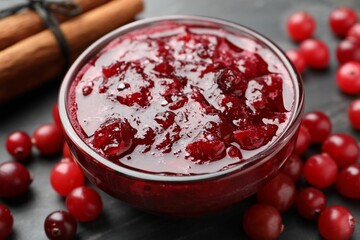 Tasty cranberry sauce in bowl, berries and cinnamon sticks on black table, closeup