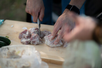 person is preparing raw meat for an outdoor meal. They are using a knife to cut the slices on a wooden cutting board
