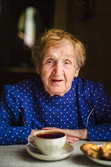 An elderly woman drinks tea at home with some pastries, enjoying a quiet moment in her cozy kitchen.