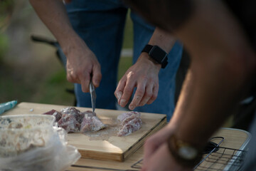 person is preparing raw meat for an outdoor meal. They are using a knife to cut the slices on a wooden cutting board