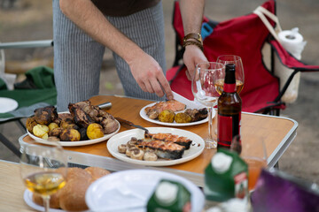 person is serving grilled meat and potatoes on a plate outside. The plate is on a table with other food, including bread and wine