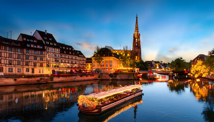 Fototapeta premium As twilight descends, a charming boat decorated with lanterns floats gracefully on the serene waters of Strasbourg's canals, surrounded by historic architecture and twinkling lights