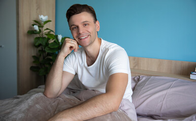 Smiling man in a white t-shirt sitting on a bed with a cozy blanket, modern bedroom decor with blue walls and a green potted plant in the background, creating a relaxed morning vibe.