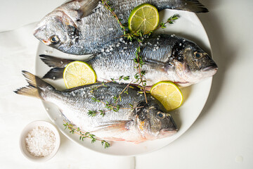 Three raw fish dorado with lime and lemon with herbs and salt and pepper on a white plate on a light table