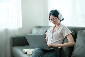 A woman wearing headphones and glasses is sitting on a couch and using a laptop
