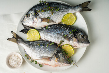 Three raw fish dorado with lime and lemon with herbs and salt and pepper on a white plate on a light table