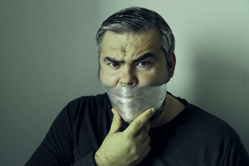 middle aged business man with transparent hand stretch foil tape over his mouth standing against a grey white wall background. scared Male with tape over lips showing no hope emotion. no talking