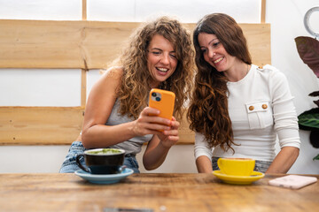 Two friends laughing at a smartphone while sitting at a coffee shop