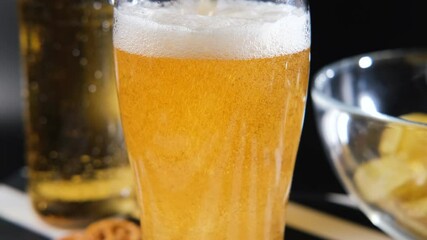 Pouring a cold beer from a bottle into a glass, creating a frothy head and bubbles rising. Nearby, a bowl filled with crispy potato chips and salty pretzels
