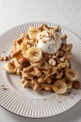 Homemade waffles stack with ice cream, bananas and nuts on white plate close up