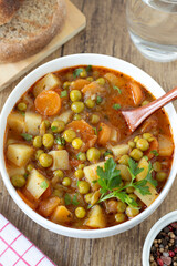 Meatless homemade vegetable stew with potato, carrot, and green peas in bowl with rustic bread on wooden table. Top view.