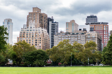 View of buildings surrounding Central Park, NYC, with lush greenery and a vibrant lawn in the foreground under a cloudy sky.