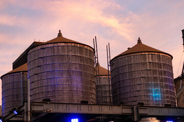 Three cylindrical wooden silos with metal roofs, illuminated by blue light accents, stand against a warm, pastel sunset sky.
