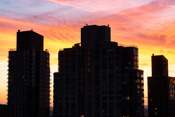 New York City rooftop view with silhouetted buildings against a vivid sunset sky in hues of orange, pink, and yellow.
