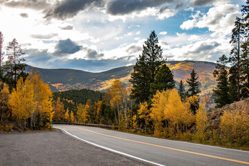 A scenic winding road in the mountains surrounded by vibrant autumn foliage and evergreen trees under a partly cloudy sky.
