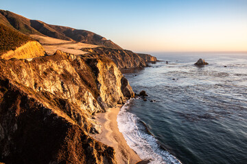 Golden coastal cliffs in Big Sur, California, with waves lapping at a sandy beach and a sunset horizon.
