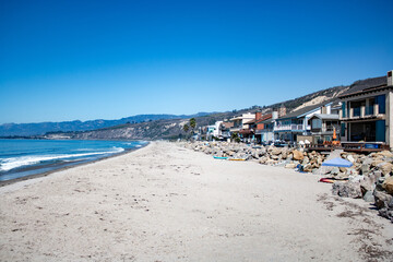 Beachfront homes along the California coast with a sandy shoreline and scenic mountain backdrop under clear skies.
