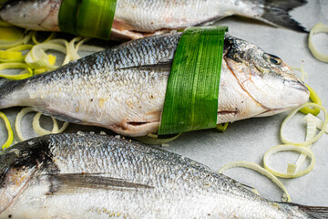 Raw, fresh dorado fish on baking paper wrapped in leek leaves with onions cut into rings.