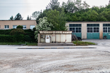 A forsaken gas station stands in an industrial complex, surrounded by overgrown greenery and aged buildings