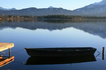 Fischerboot am See, links ein Holzsteg, im Hintergrund Wald, Berge, Himmel, Wasserspiegelung. Hopfen am See bei Füssen, Allgäu, Bayern