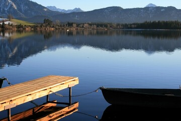 Fischerboot am See, links ein Holzsteg, im Hintergrund Wald, Berge, Himmel, Wasserspiegelung. Hopfen am See bei Füssen, Allgäu, Bayern
